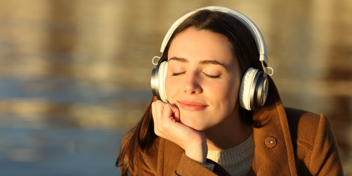 A young woman closing her eyes and leaning her head on her hand as she listens to music through her headphones. She is resting by a lake